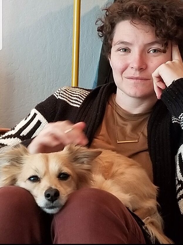 Image shows Bonnie, a woman with curly red hair, sitting with a small golden colored dog in her lap. Bonnie is resting her head on her hand and has a slight smile on her face.