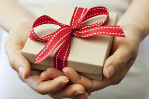 Image of two hands holding a small beige gift box. The box is wrapped with red and white spotted ribbon with a bow.