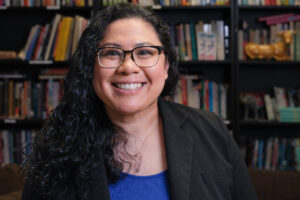 Person with long curly hair and glasses is wearing a dark colored jacket outside a blue shirt.